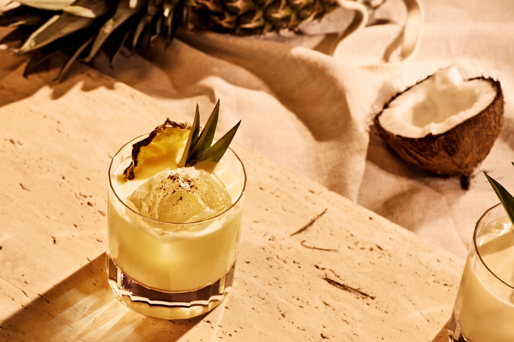 Rommicocktail lasissa, taustalla puolikas kookospähkinä ja ananaksen lehtiä.