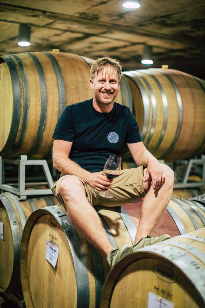 Fabian Schwarz istuu viinitynnyreiden päällä hymyillen viinilasi kädessään