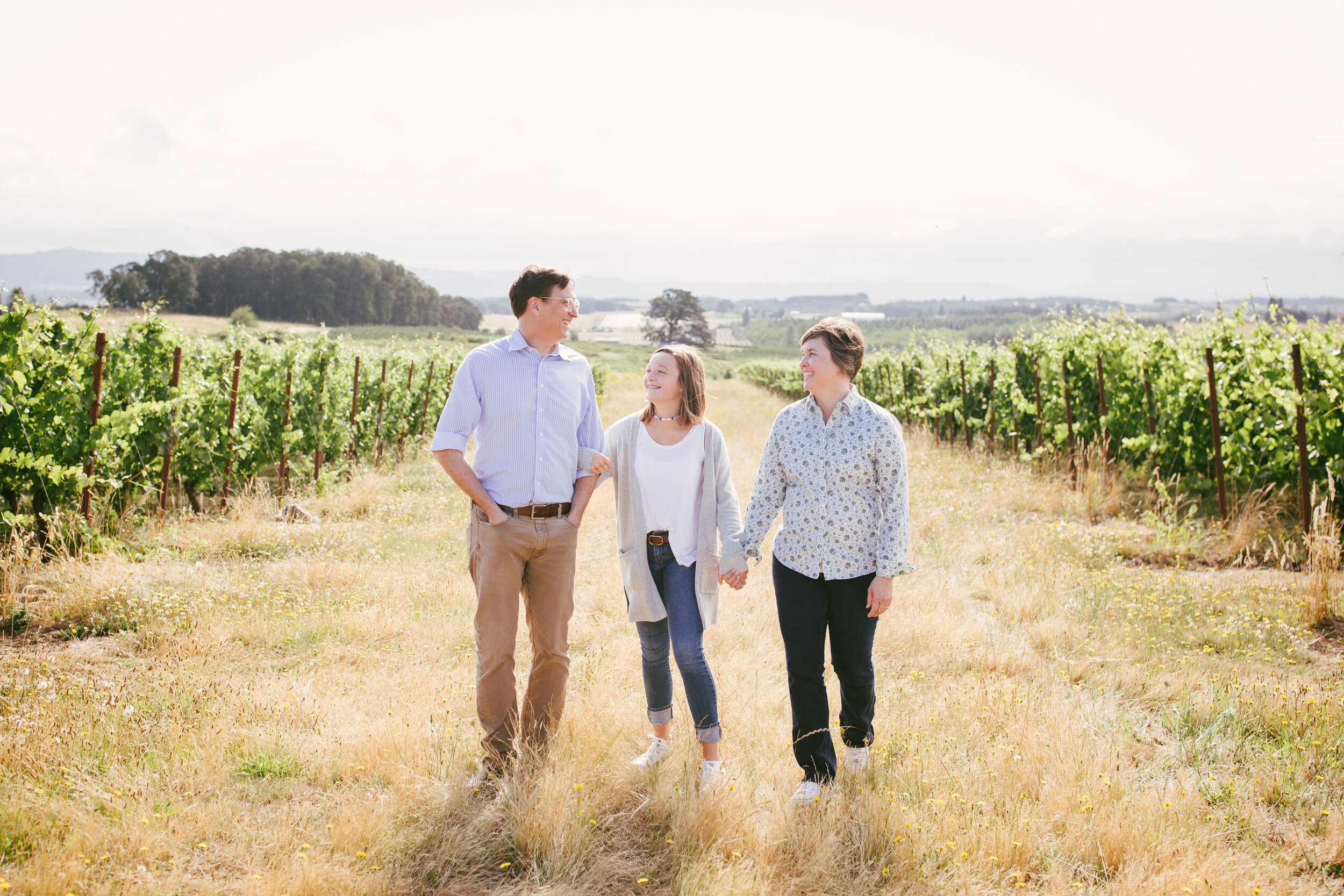 Perhe kävelemässä maalaismaisemassa viiniviljelmien keskellä käsi kädessä.