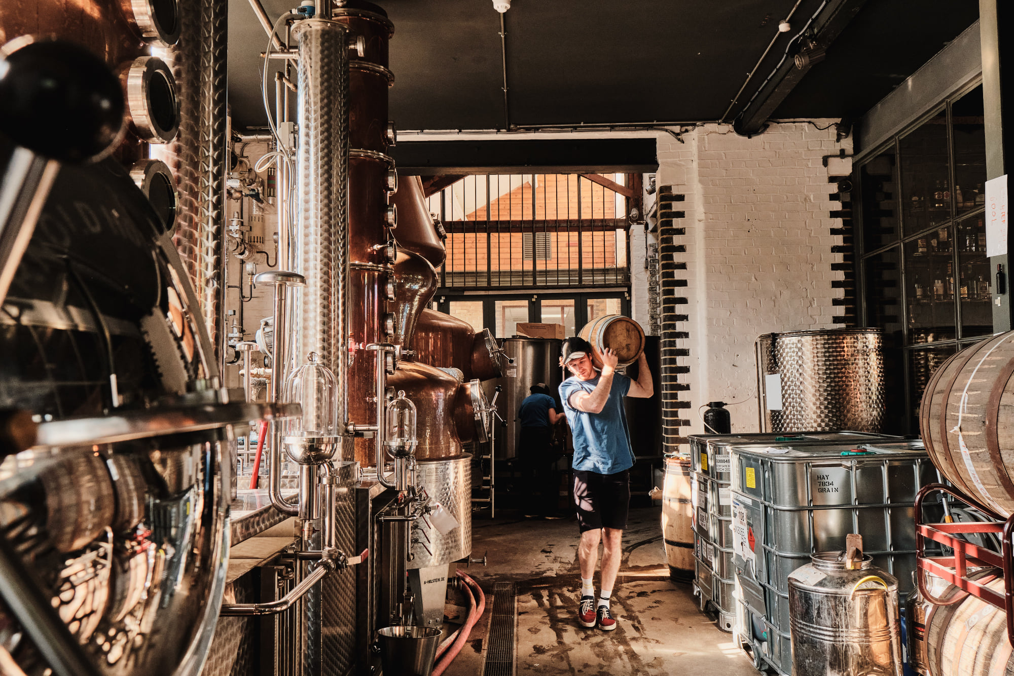 Tislaamotuotannon laitteita tislaamosssa sekä henkilö kantamassa keskikokoista tai pientä tynnyriä olallaan.