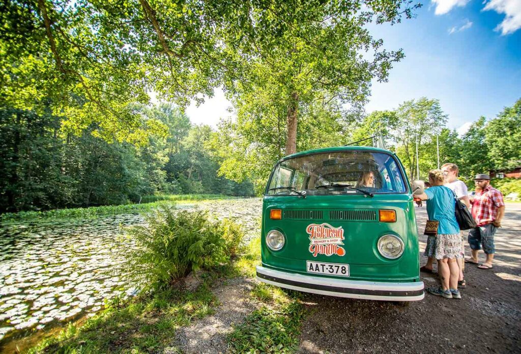 Kaunis vehreä ja kesäinen jokimaisema, ja joen äärelle parkkeerattu vanha vihreä pakettiauto, jonka ääreen on kerääntynyt ihmisiä.