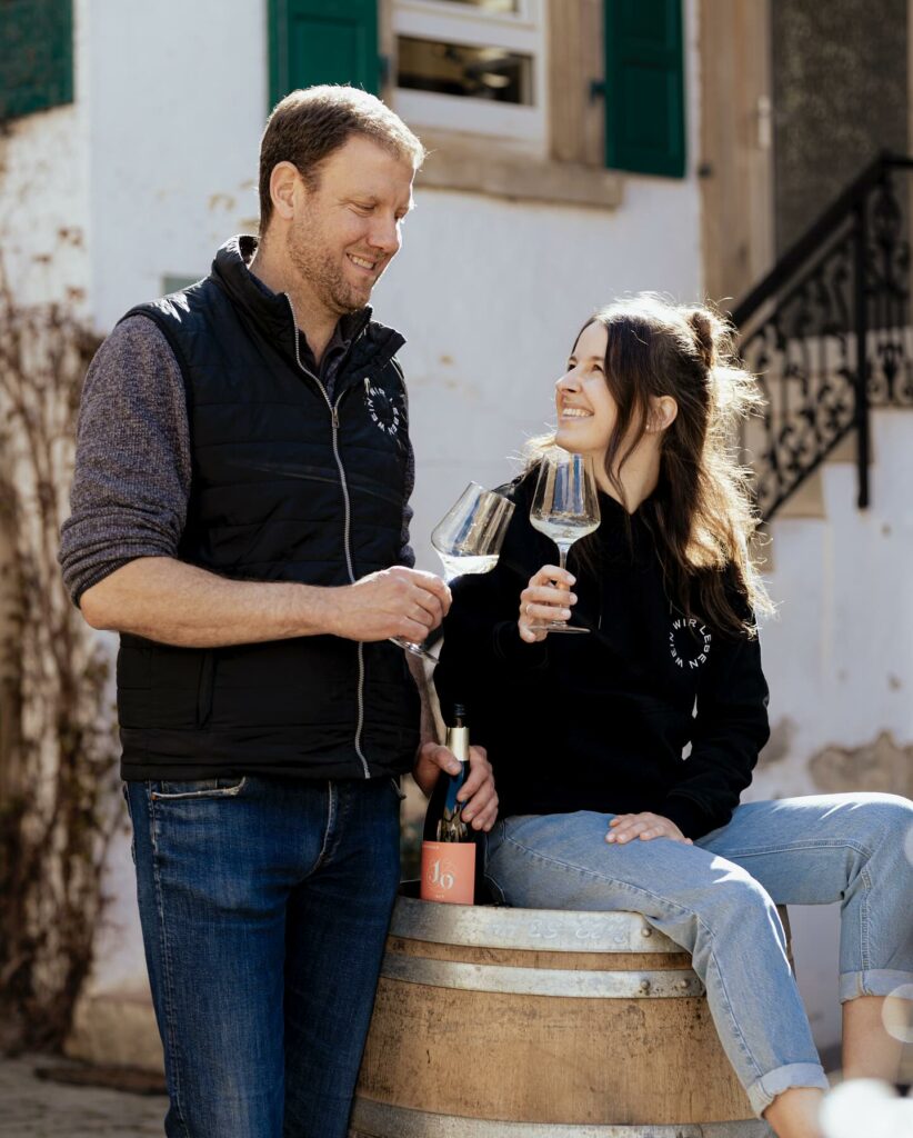 Johanningerin Oliver Herzer ja Lara Haas skoolaamassa viinileaseilla ulkona talon edustalla. Lara istuu viinitynnyrin päällä.