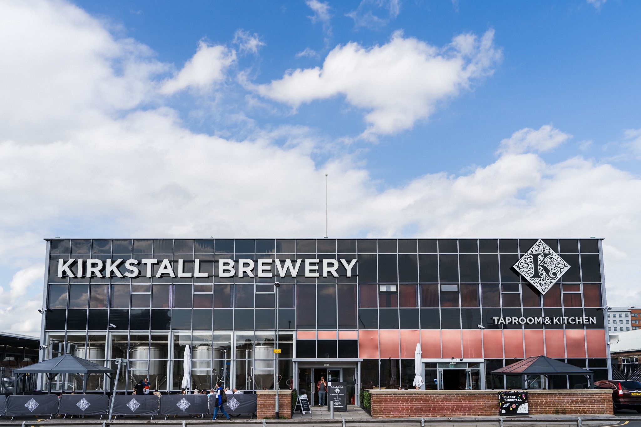 Kuva laatikonmallisests rakennuksesta, jossa lukee isolla Kirkstall Brewery. Ikkunoista näkee läpi hieman sisällä olevia terästankkeja. Ulkona on terassi.