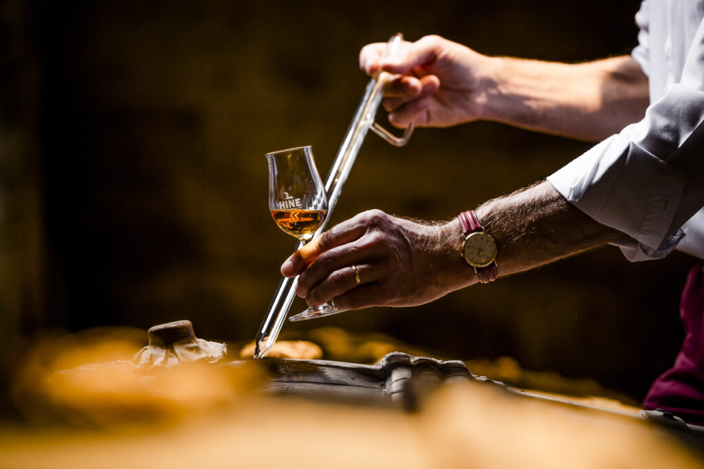 Konjakkilasi ja tynnyri sekä kädet, jotka ottavat välineellä konjakkia tynnyristä lasiin.