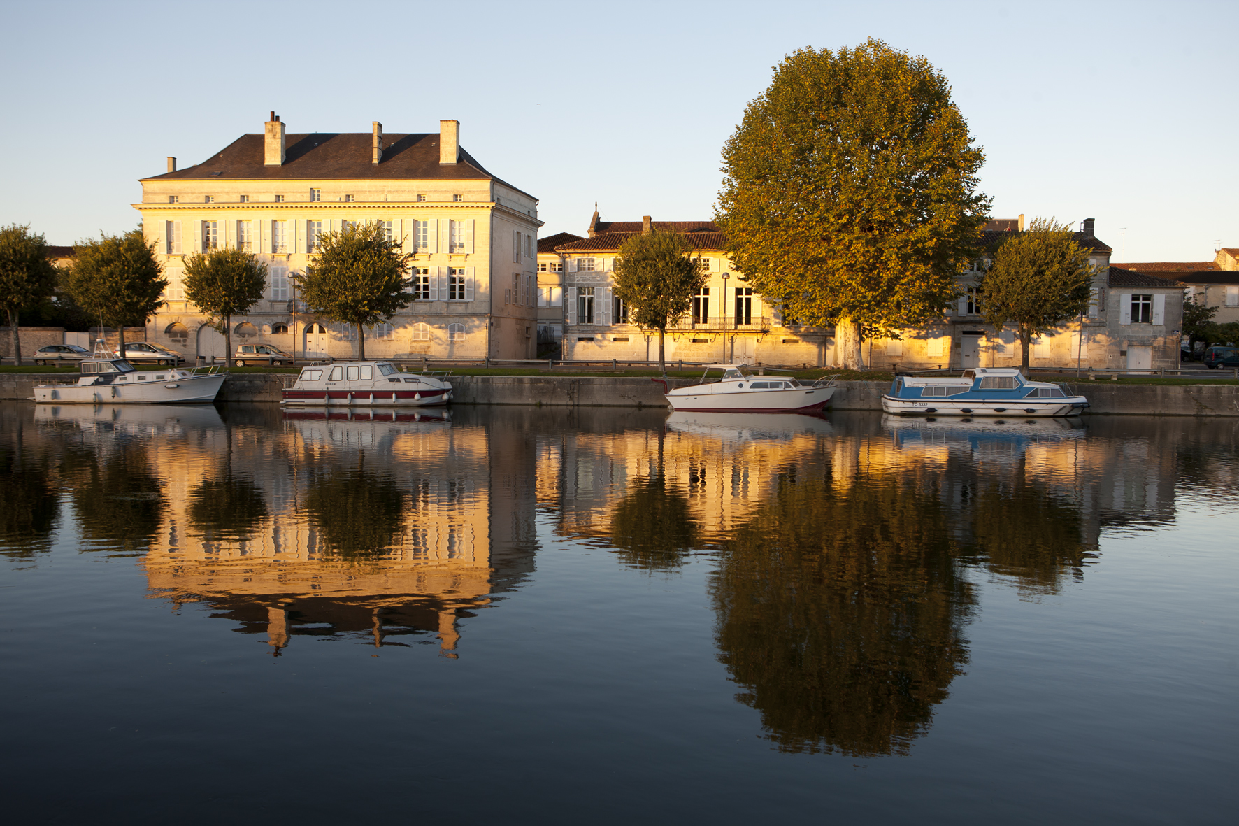 Hine Cognacin talo joen rannalla auringonlaskussa. Talo, puu ja veneet heijastuvat vedenpinnasta.