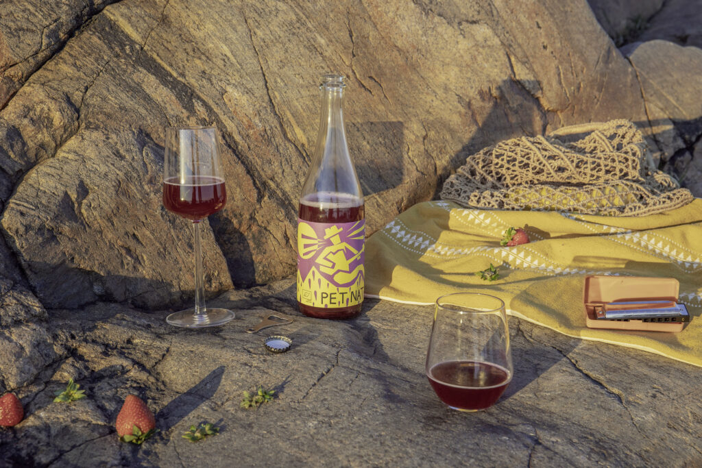 Noita Winery Pet Nat -pullo auringossa kalliolla piknikviltin vieressä. Kahdessa erilaisessa lasissa kirkasta punaista natuviiniä  kaadettuna pullosta. Mansikoita maassa.