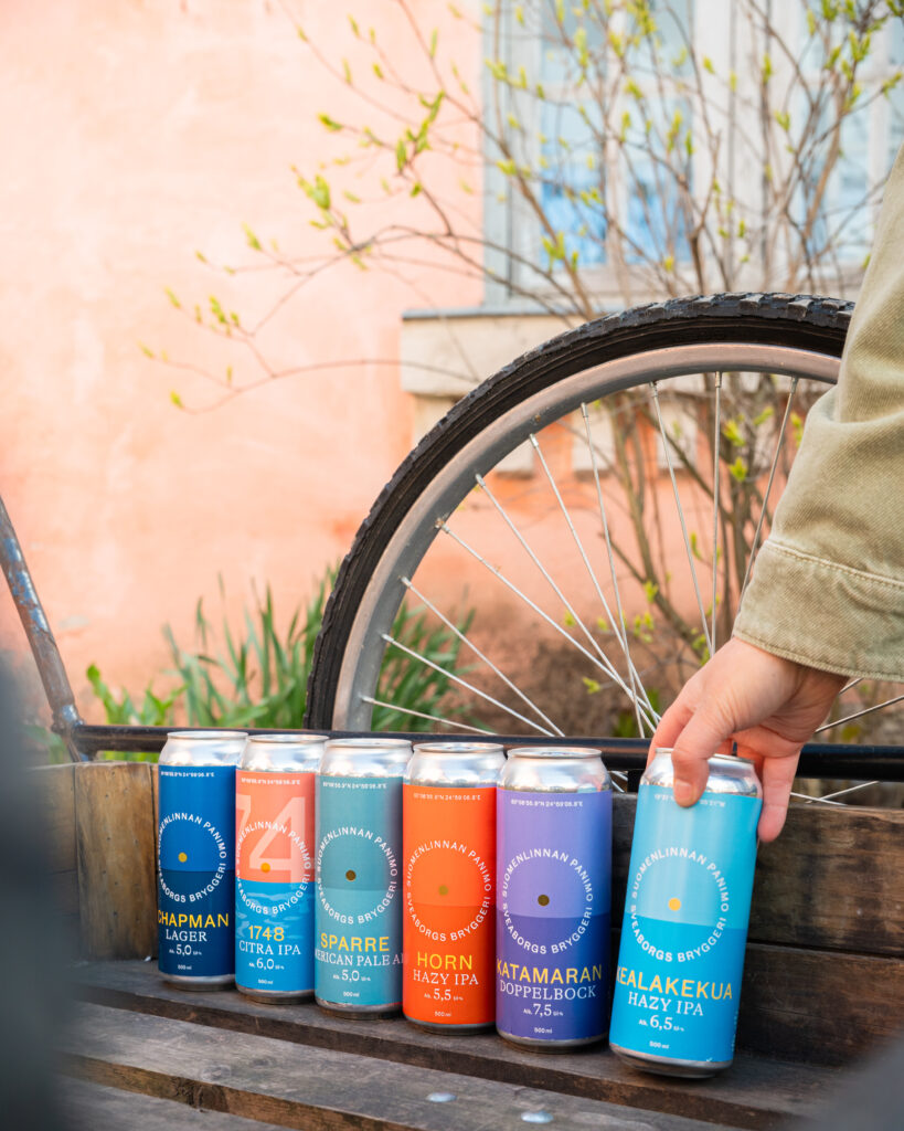 Kuusi erilaista värikästä Suomenlinnan Panimon tölkkiä rivissä, kuvattuna kärryissä Suomenlinnassa.